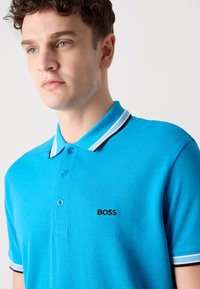 Tirkizna polo majica od teksturirane tkanine, s placketom na tri dugmeta i kontrastnim prugastim kragnom. Sadrži crni logo "BOSS" na prsima.
