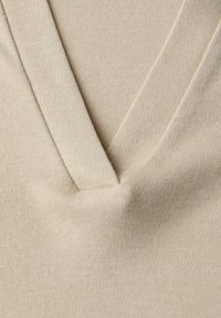 Gros plan sur un tissu en maille beige avec un détail d'encolure en V, montrant une couture fine et une texture.