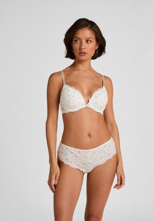 Vrouw met kort donker haar, die een witte bloemenkant beha en bijpassend kanten ondergoed draagt, staand tegen een effen grijze achtergrond.