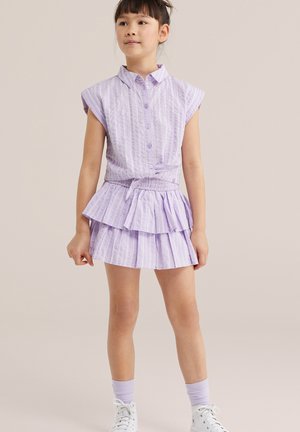 Niña joven de pie, vistiendo una camisa sin mangas de rayas lavanda con botones y una falda plisada a juego, con zapatillas blancas y calcetas lavanda.