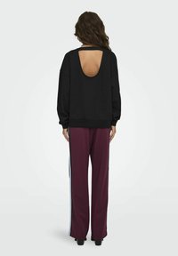 Zwarte sweater met een grote uitsnijding aan de achterkant, gecombineerd met bordeauxrode broek met zijstrepen in lichtblauw. Het materiaal lijkt zacht en comfortabel.
