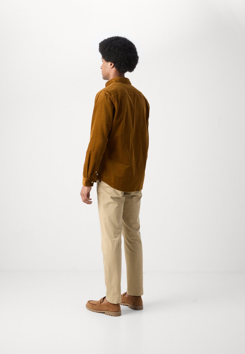 Braunes Cordhemd, beige Hose und braune Schuhe. Modell von hinten gesehen, mit lockigem Haar und einem neutralen Hintergrund.