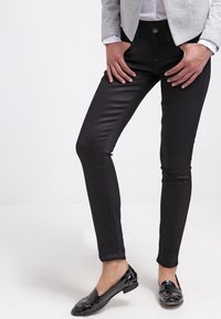 Pantalones negros ajustados con una textura suave, que cuentan con un cierre de botón y presillas para cinturón. Combinados con mocasines negros de charol y una blazer gris.