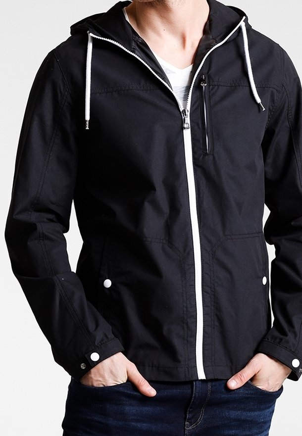 Veste à capuche noire avec fermeture éclair blanche et cordons portée par une personne les mains dans les poches, associée à un jean bleu foncé.
