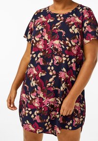 Kvinna iklädd en kortärmad marinblå klänning med ett abstrakt blommönster i rosa, beige och brunt, stående framför en vit bakgrund.