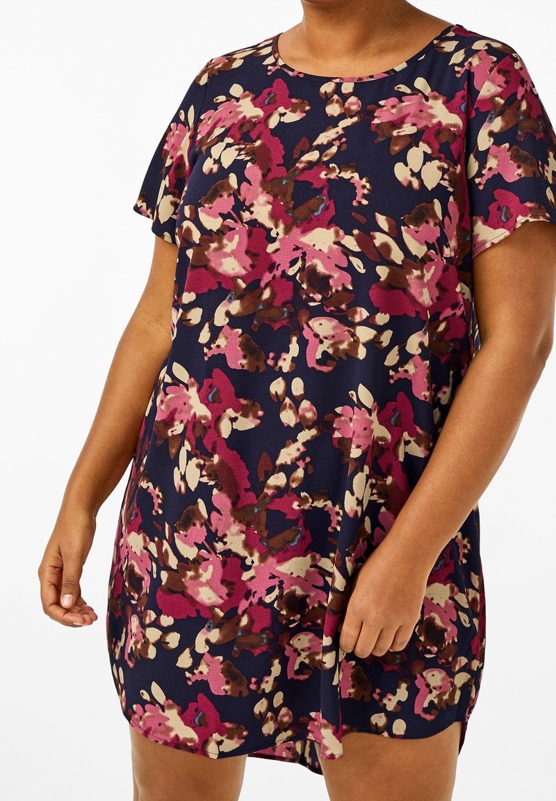 Kvinna iklädd en kortärmad marinblå klänning med ett abstrakt blommönster i rosa, beige och brunt, stående framför en vit bakgrund.