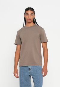 Young man with long braided hair wearing a plain brown short-sleeve t-shirt and light blue jeans standing against a white background.