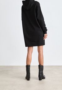 Vestido sudadera negro de gran tamaño con capucha, que presenta puños acanalados y un dobladillo recto; combinado con botas de tacón altas negras hasta la rodilla.