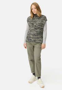 Gilet en tricot dans des tons noirs et beiges avec un col haut. Associé à un pantalon vert olive. Le mannequin porte des baskets blanches.