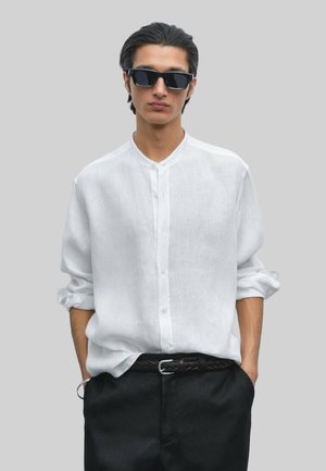 White linen shirt with a mandarin collar and button closure, paired with dark trousers and a woven black belt, showcased against a gray background.