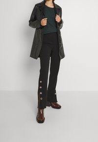 Manteau long en cuir noir brillant, pull vert texturé, pantalon noir sur mesure avec des fentes latérales et des accents de boutons, chaussures à talons marron.