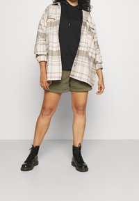 Camisa de cuadros beige oversize con detalles en negro, superpuesta sobre una sudadera negra, combinada con pantalones cortos verde oliva y botas de combate negras con cordones.