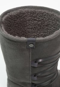 Bottine en suede gris avec doublure en textile pelucheux, haut arrondi, semelle en caoutchouc plate, détail de lacets sur le côté et étiquette de marque au niveau de la cheville.