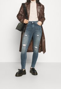 Trench de cuero marrón sobre una blusa beige, combinado con jeans azules desgastados con rotos. Zapatos de plataforma negros y un bolso negro pequeño.