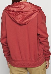Červená mikina s kapucí na šňůrku, s žebrovanou texturou a elastickými manžetami. Měkká tkanina s volným stříhem a nápadně staženým spodním lemem.
