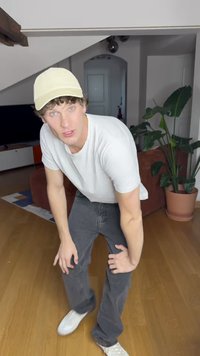 Vit t-shirt, ljusgul keps, mörkgrå jeans och vita sneakers. Miljön inkluderar en brun soffa och en krukväxt.
