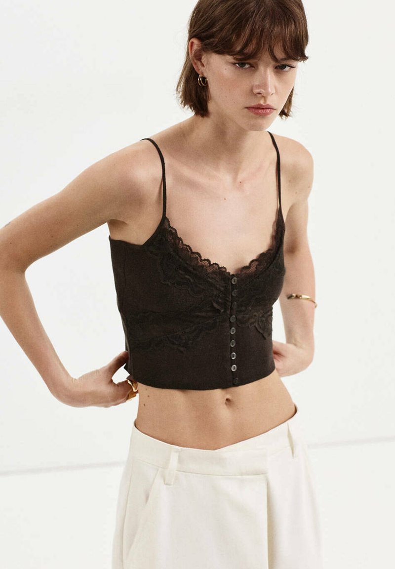 Femme portant un caraco court noir à bordure en dentelle avec des boutons et un pantalon blanc taille haute sur un fond uni.