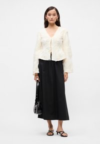 Biely textúrovaný blúzka s výstrihom do V a dlhými rukávmi, kombinovaná s čiernou maxi sukňou, čiernymi šlapkami a malou čiernou taškou.