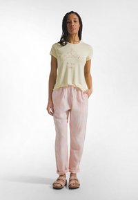 Lichtgeel T-shirt met korte mouwen en een minimalistisch grafisch ontwerp. Losvallende lichtroze linnen broek met een elastische tailleband. Bruine sandalen.