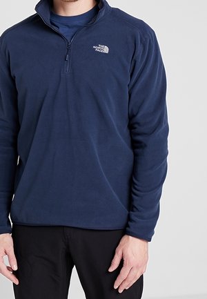 Hombre con un forro polar azul marino con cremallera hasta el pecho y el logo de The North Face en el pecho, y pantalones negros sobre un fondo gris claro.