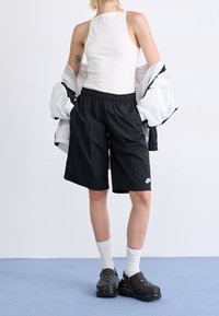 Person trägt ein weißes ärmelloses Oberteil, schwarze Nike-Shorts, weiße Socken, schwarze Clogs und eine weiße Jacke, die über die Schultern gehängt ist.