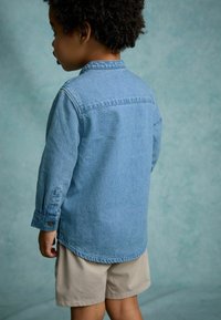 Camisa de mezclilla, azul claro, de manga larga, con puños abotonados y dobladillo redondeado. Llevada con pantalones cortos beige, mostrando un estilo casual en capas.
