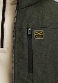 Olive green jacket with a textured surface, featuring a black zipper, a prominent brand label, and a contrasting cream interior.