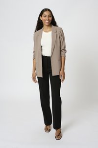 Femme aux longs cheveux foncés portant un blazer beige, un top blanc, un pantalon noir et des talons ouverts noirs, debout et souriante devant un fond uni.