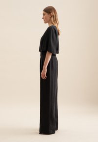 Czarny lniany kombinezon z krótkimi rękawami, szerokimi nogawkami i bocznymi kieszeniami. Gładka textura i minimalistyczna, nowoczesna sylwetka.