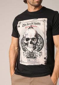 T-shirt noir à manches courtes avec un graphique de crâne, un design de carte à jouer, et le texte "L'As de Pique" en rouge et blanc.