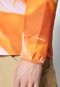 Orange vindjakkeærme med elastisk manchet, glat tekstur og trykt abstrakt design. "WINDWALL"-logoet er synligt nær manchetten.