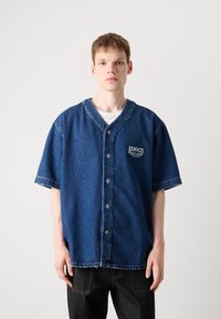 Blå denim kortermet skjorte med knappfeste foran og v-hals, med en liten logo på brystet og frynsete kanter.
