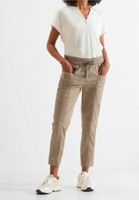 Pantalones jogger beige con cintura elástica y cordón. Dos bolsillos laterales, pierna ajustada, y combinados con una blusa blanca de cuello en V y zapatillas deportivas.