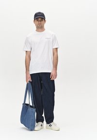 Valge puuvillane T-särk logoga, tumedad denimpüksid, sinine denimpoolkott ja valged tennisetossud, millel on tumesinised aktsendid, kandes seisvat meessoost figuuri.