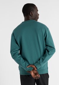 Sudadera turquesa con mangas largas, puños acanalados y cuello redondo. Textura suave, diseño sencillo sin patrones o detalles visibles.