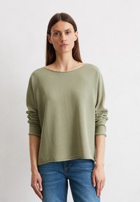 Hellgrüner, weicher Strickpullover mit weitem Bootsausschnitt und langen Ärmeln. Ribbenbündchen und Saum, getragen mit blauen Jeans vor einem neutralen Hintergrund.