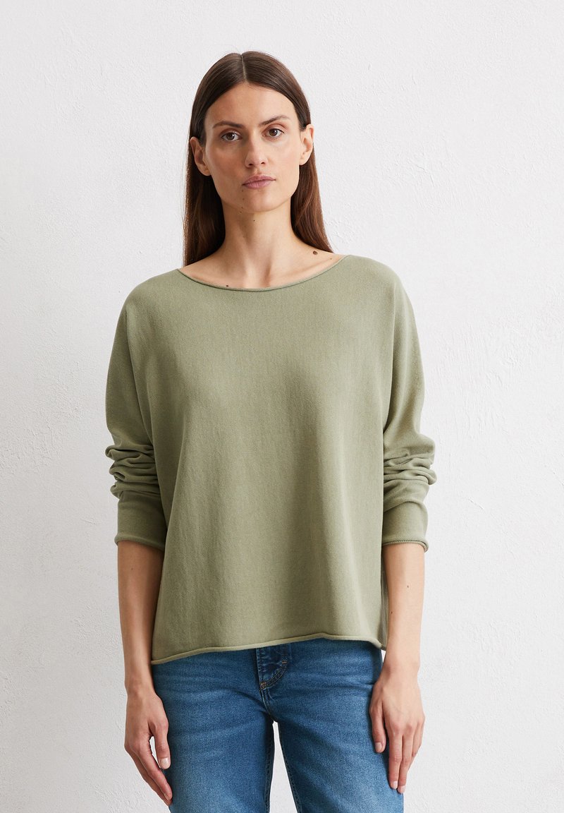 Hellgrüner, weicher Strickpullover mit weitem Bootsausschnitt und langen Ärmeln. Ribbenbündchen und Saum, getragen mit blauen Jeans vor einem neutralen Hintergrund.