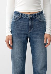 Persona vistiendo jeans de mezclilla azul de tiro medio y una camiseta de tirantes larga blanca de canalé, de pie con las manos relajadas a los lados.