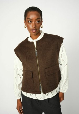 Bruine wollen vest met een hoge kraag, ritssluiting aan de voorkant, twee voorzakken en korte mouwen, gelaagd over een witte textuurblouse met uitsnijdingen.