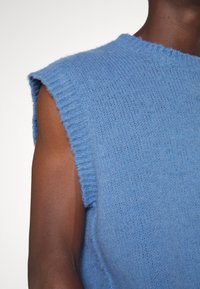 Gros plan sur le bras supérieur et l'épaule d'une personne portant un pull bleu sans manches en tricot avec des bords côtes.