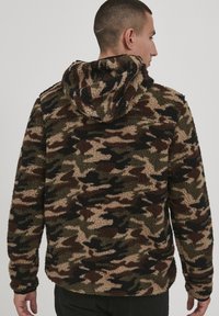 Fleece hoodie in camouflage pattern with shades of brown, green, and black. Features a textured surface and a hood. Back view shown.