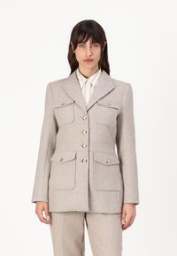 Beige houndstooth-blazer med to brystlommer og knapper, med notched lapel og skreddersydd passform. Stoffets tekstur virker jevn og strukturert.