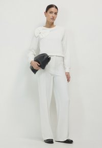 Weißer Pullover mit strukturiertem Schleifendetail an der Schulter, kombiniert mit hoch geschnittenen weißen weit geschnittenen Hosen und einer schwarzen Clutch.