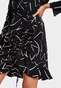 Svart kjol med vita abstrakta mönster, med rynkdetaljer längs sidan och en volangkant, tillverkad av ett lättviktsmaterial.