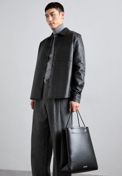 Svart läderjacka med knappfront, grå polotröja underneath, och grå ullbyxor. Håller en svart läderhandväska med logotyp.