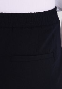 Zwarte broek met een elastische tailleband. Beschikt over een achterzak met een eenvoudige klep. De stof lijkt glad en licht gestructureerd.