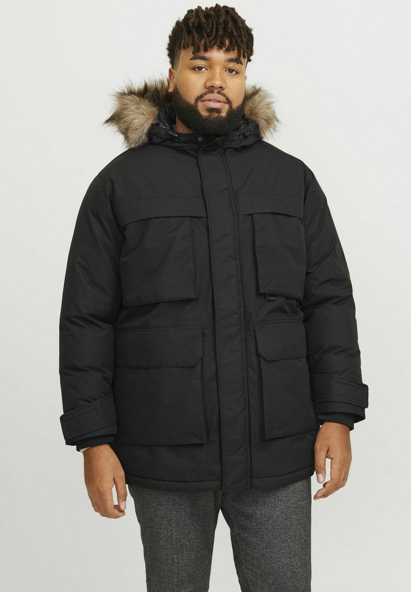 Chaqueta acolchada negra con capucha forrada de piel, múltiples bolsillos frontales, cierre de cremallera y puños elásticos, fabricada con un material liso y resistente a la intemperie.