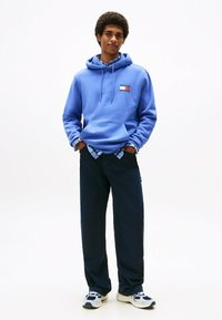 Blaues Kapuzensweatshirt mit einem kleinen rechteckigen Logo, kombiniert mit dunklen Hosen und weißen Sneakern mit blauen Akzenten. Lässiger, entspannter Schnitt.