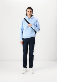 Lys blå hettegenser med lomme foran, kombinert med mørke jeans og hvite joggesko. Modellen holder et svart bagstropp over kroppen.