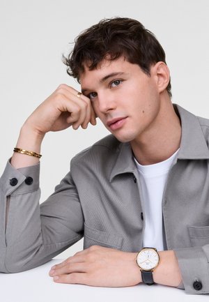Camisa de botones gris con botones negros, de textura pesada, llevada sobre una camiseta blanca. Reloj dorado con carátula blanca y correa de cuero negro en la muñeca.
